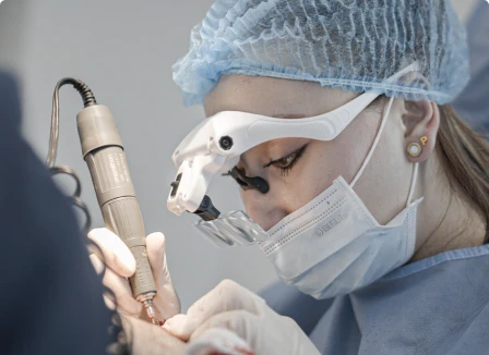 Un primer plano de una cirujana especialista en implante capilar, que lleva gorro quirúrgico, mascarilla y gafas de aumento quirúrgicas con iluminación, mientras utiliza un instrumento de precisión de mano para realizar una implantación folicular FUE.